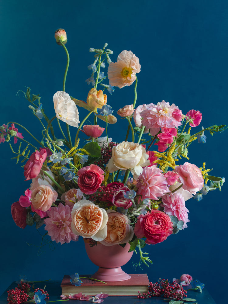 The Pink Kiss Poster features a vibrant floral arrangement with roses, poppies, and delphiniums in romantic shades of pink, peach, and cream. Set against a bold blue backdrop in a soft pink vase, it embodies elegant still life photography.