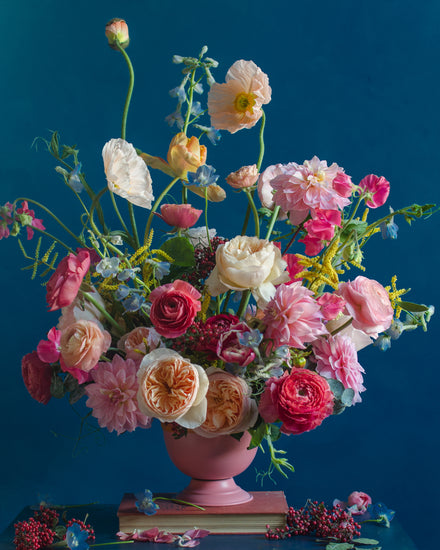 The Pink Kiss Poster captures a vibrant bouquet of romantic flowers including roses, poppies, and peonies in pink, peach, and cream hues. Elegantly arranged in a pink vase against a rich blue backdrop, this still life photograph evokes timeless beauty.