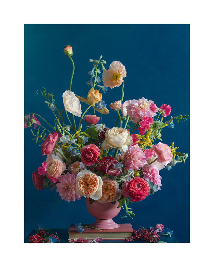 The Pink Kiss Poster features a vibrant floral arrangement of pink, peach, and white blooms in a pink vase set against a deep blue backdrop. Roses, poppies, and dahlias gracefully reach upward with berries artfully scattered below, embodying an elegant style in still life photography.