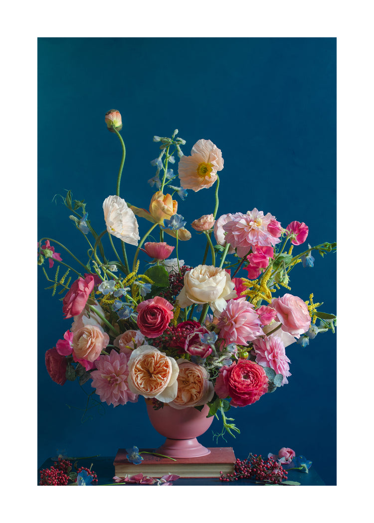 The Pink Kiss Poster showcases an elegant floral arrangement in a pink vase set against a deep blue background. It features roses, poppies, and wildflowers in pink, red, orange, and white hues with some blooms artfully scattered at the base, evoking romantic flowers.