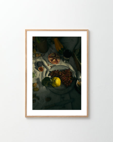 The Gloomy Picnic III Poster displays a still life with a plate featuring lemon and grapes, sliced pomegranate, figs, and red wine on white cloth. Fig leaves add depth, complemented by scattered utensils and various fruits.