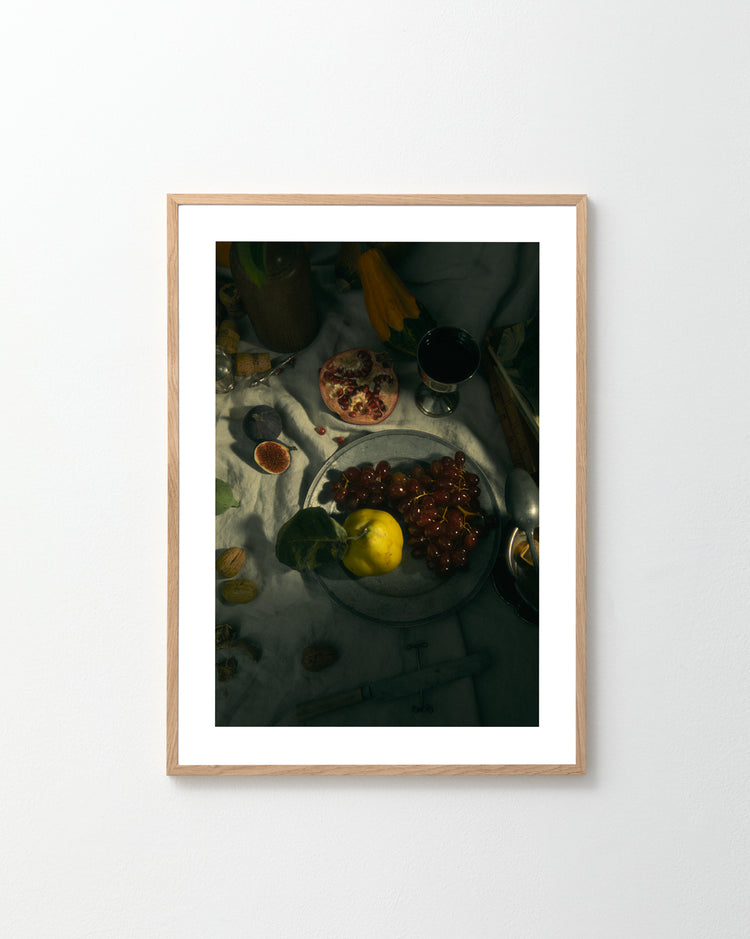 The Gloomy Picnic III Poster displays a still life with a plate featuring lemon and grapes, sliced pomegranate, figs, and red wine on white cloth. Fig leaves add depth, complemented by scattered utensils and various fruits.