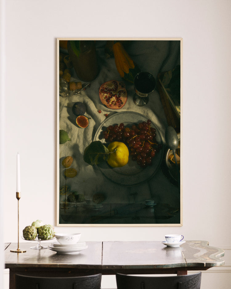 A photo of a dining area with a wooden table featuring a candle, vase, and teacup. Above it hangs the Gloomy Picnic III poster, a large still-life scene reminiscent of an Adam and Eve tableau with grapes, pomegranate, and yellow apple.