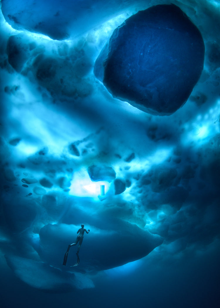A diver swimming in the deep blue ocean underneath a surface made from ice.