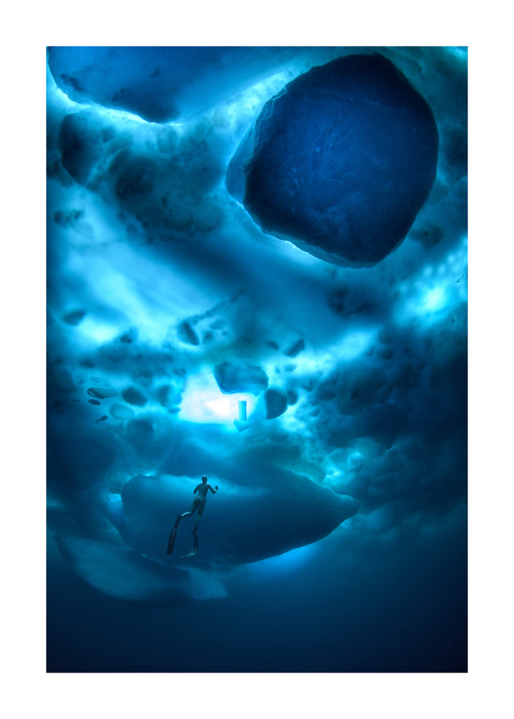 A diver swimming in the deep blue ocean underneath a surface made from ice.