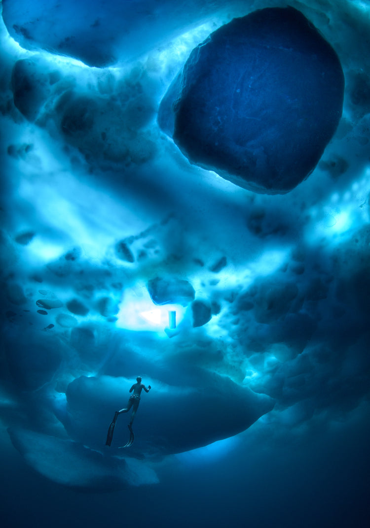 A diver swimming in the deep blue ocean underneath a surface made from ice.