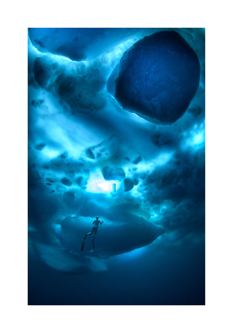 A diver swimming in the deep blue ocean underneath a surface made from ice.