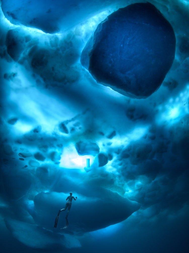 A diver swimming in the deep blue ocean underneath a surface made from ice.
