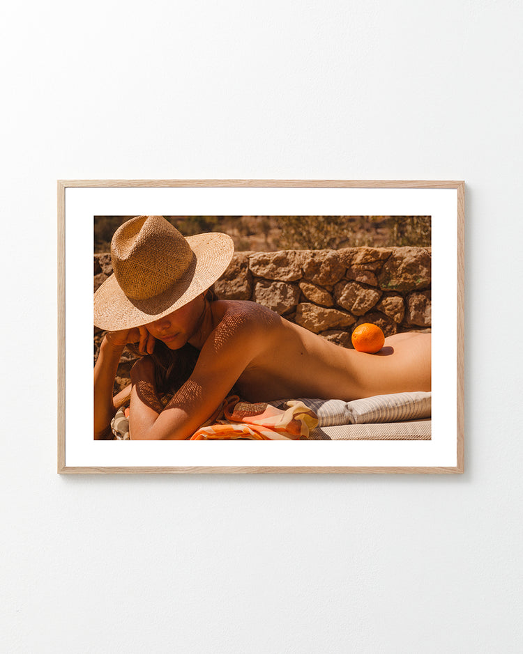 Naked woman on a sun bed wearing a hat and balancing an orange on her back.