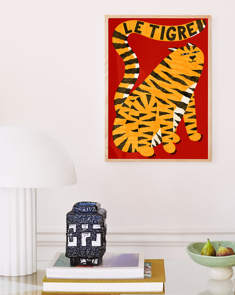 The Le Tigre Red Poster by Manne Jalilian, featuring a stylized orange tiger on a striking red background, is framed and hung on a white wall. Below it sits a modern white lamp with a dome shade on a table, alongside stacked books, a small ceramic piece, and nearby bowl of green apples.