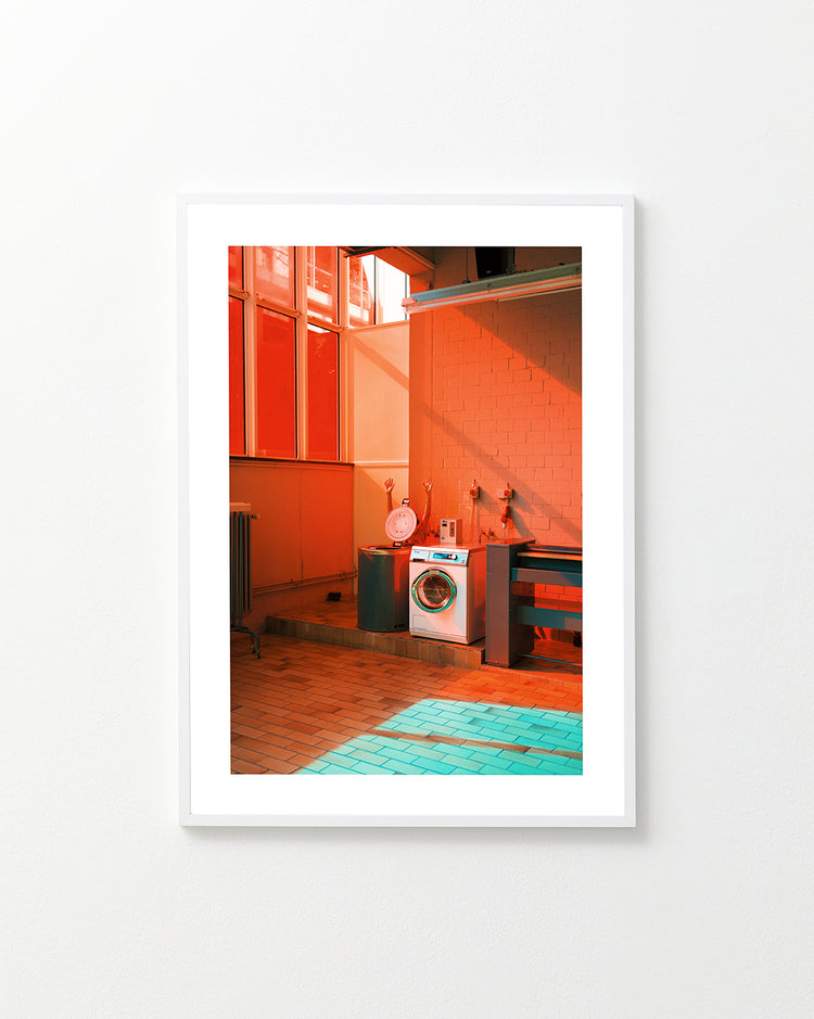 The Laundry Day Poster by Delfina Carmona features a laundromat scene with a washing machine and chair in striking red and turquoise lighting. Sunlight casts geometric shadows on the floor, creating a modern and artistic ambiance akin to captivating wall art.