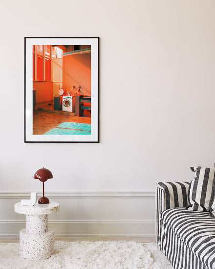 A living room features a striped sofa and a small round table with a brown lamp. A framed Laundry Day Poster by Delfina Carmona hangs on the wall, while the light wood floor is complemented by a plush white rug nearby.