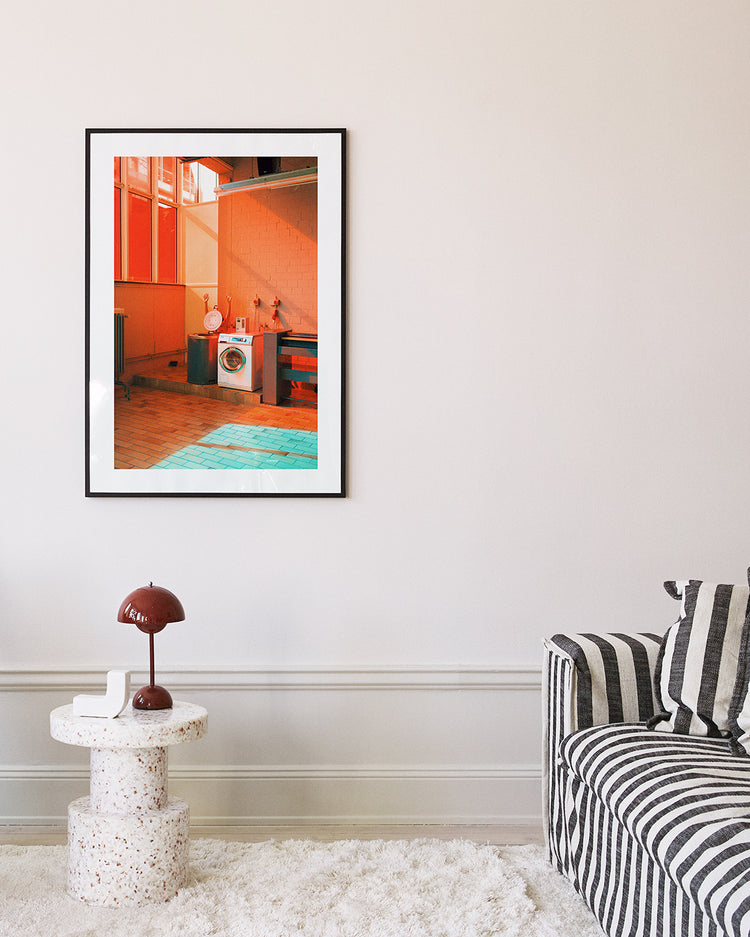 A living room features a striped sofa and a small round table with a brown lamp. A framed Laundry Day Poster by Delfina Carmona hangs on the wall, while the light wood floor is complemented by a plush white rug nearby.