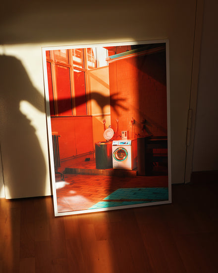 The Laundry Day Poster features Delfina Carmonas photo of a vintage decor interior with a washing machine. An intriguing shadow of an outstretched hand falls on the frame, enhanced by warm lighting, evoking a cozy laundromat feel.