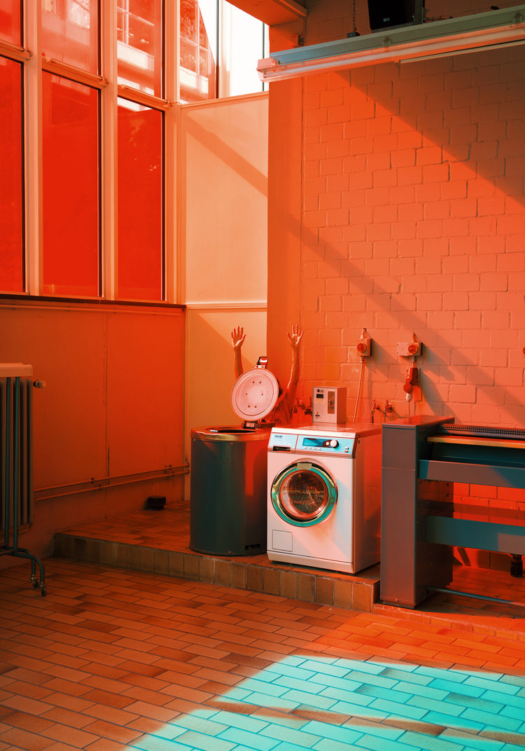 A room bathed in red-orange light, reminiscent of a Laundry Day Poster, features a washing machine and a laundry basket. Two mannequin arms whimsically emerge from the basket as sunlight and shadows dance across the brick walls and tile floor, evoking an artful laundromats charm.
