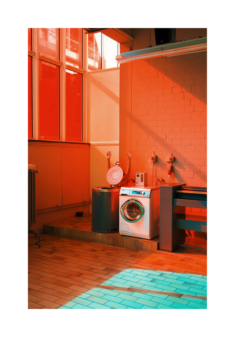 Bright red and orange hues fill a laundromat with a washing machine against a brick wall, near a basin. Light streams through large windows, casting colored shadows on the tiled floor where two mannequin arms rise from a drum—evoking the avant-garde feel of the Laundry Day Poster.