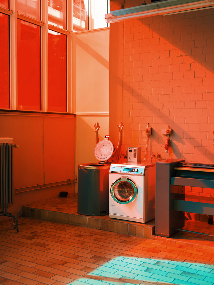 The Laundry Day Poster showcases a laundromat room with a washer and dryer against a brick wall, lit by warm orange and red hues. Mannequin arms mysteriously emerge from the dryer, casting intriguing shadows, capturing Delfina Carmonas flair for compelling visual stories.