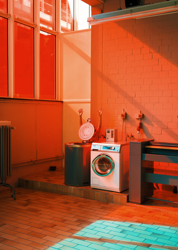 A laundromat scene with a red-orange hue features a white washing machine and a black basket with mannequin arms poking out. Sunlight through large windows creates playful shadows on the tiled floor, evoking the style of the Laundry Day Poster by Delfina Carmona.