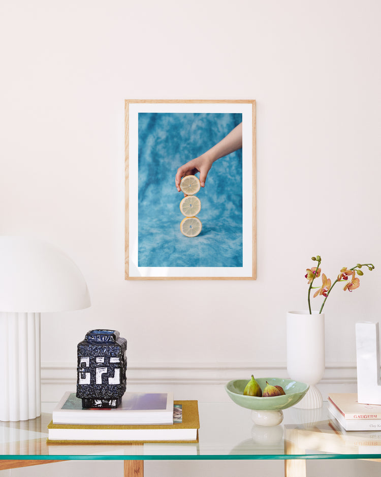 A minimalist room displays a framed Sour Poster, featuring a hand holding a lemon slice against a blue background. Below, on the glass table, are a modern lamp, decorative jar, books, a bowl with figs, and an orchid in a vase.