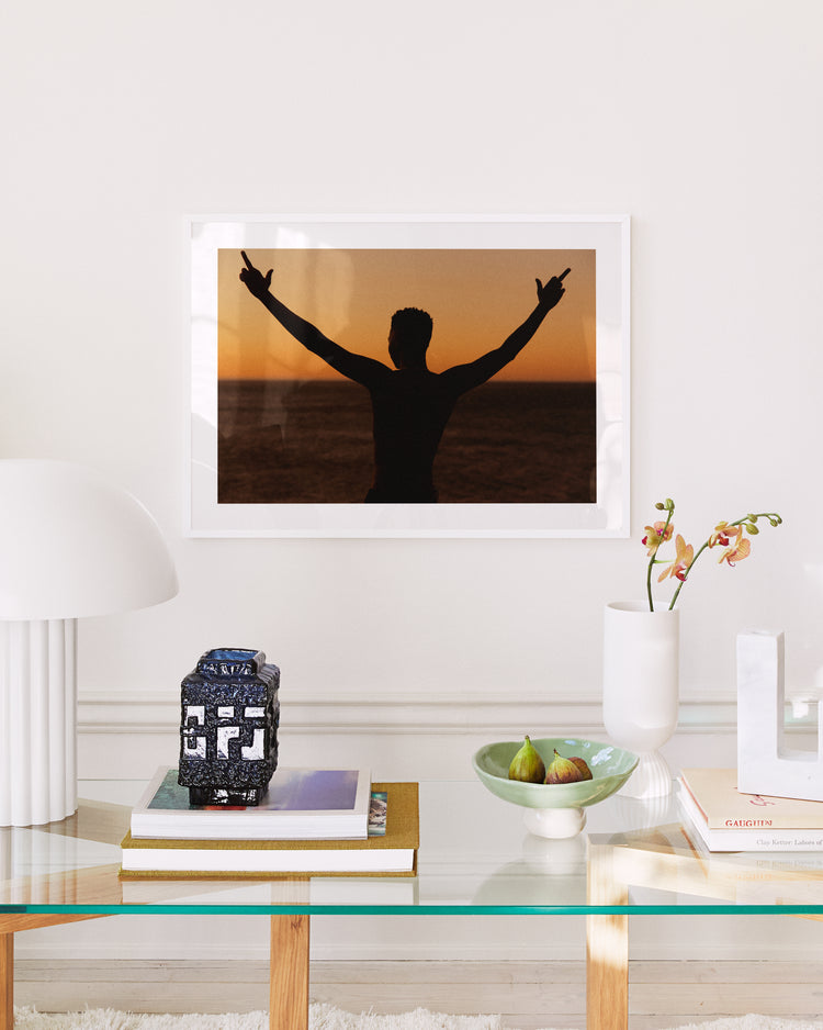 A modern living room showcases a glass table adorned with books, a decorative jar, figs in a bowl, and an orchid. Above, the My New Tinder Picture Poster captures the bold try of embracing endless possibilities against a vibrant sunset backdrop.
