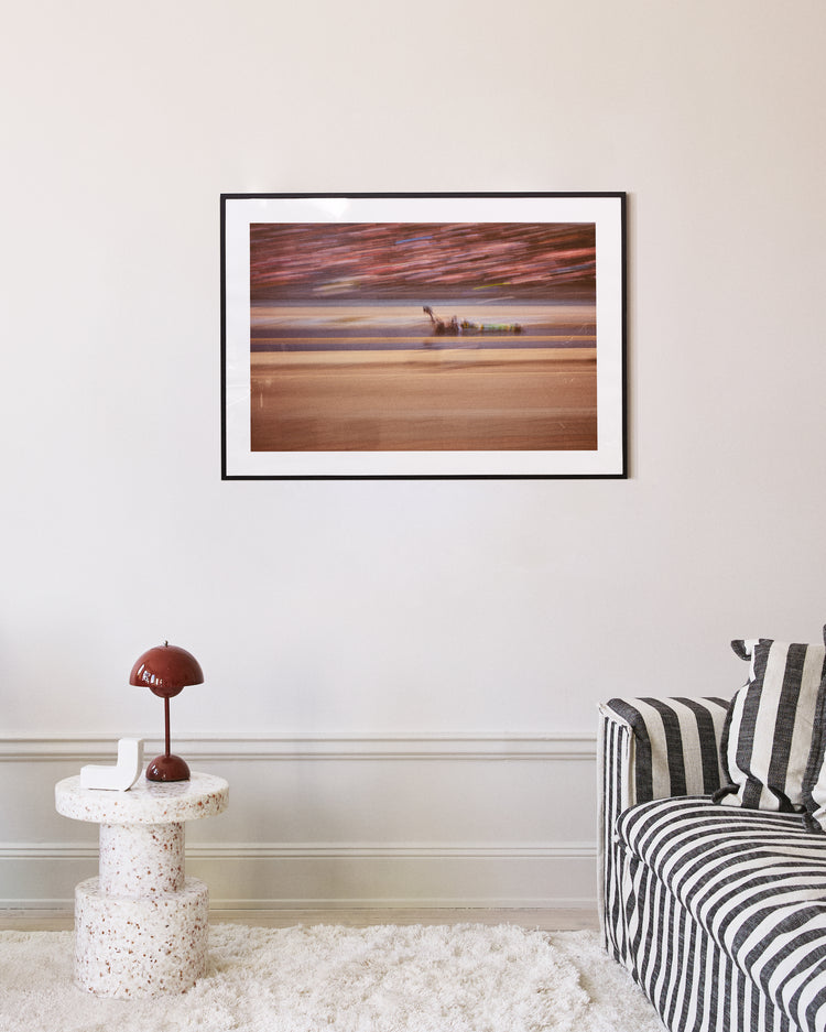 The Limitless Poster, a framed abstract beach scene, hangs on a beige wall above a modern round speckled table with a brown lamp. Nearby, a gray and white striped couch is enhanced by two accent pillows.