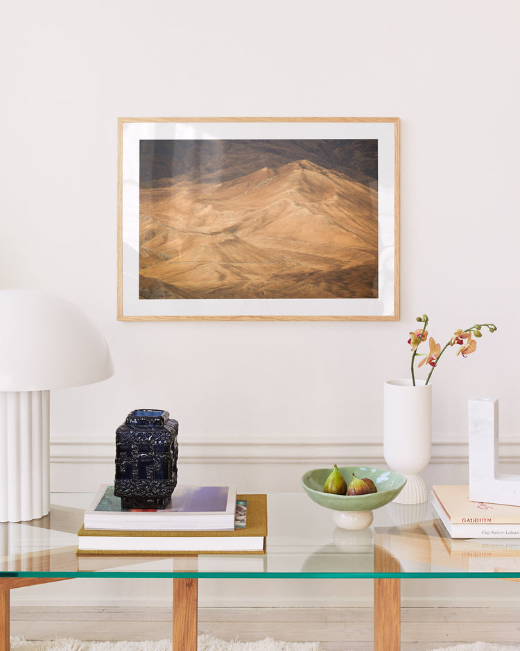 A modern living room blends nostalgia with a framed Mount Blues Poster on the wall. A glass coffee table with wooden legs holds a round lamp, black decorative box, stacked books, green bowl, and pink flowers in a white vase.