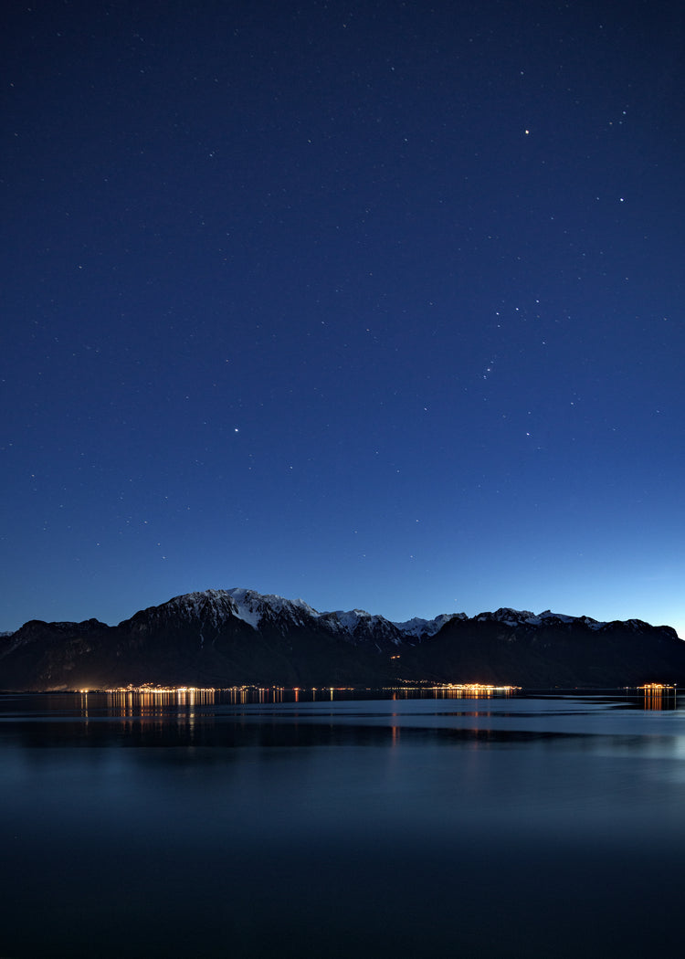 Mountain scenery, lights from a city and a lake under a star strewn sky.