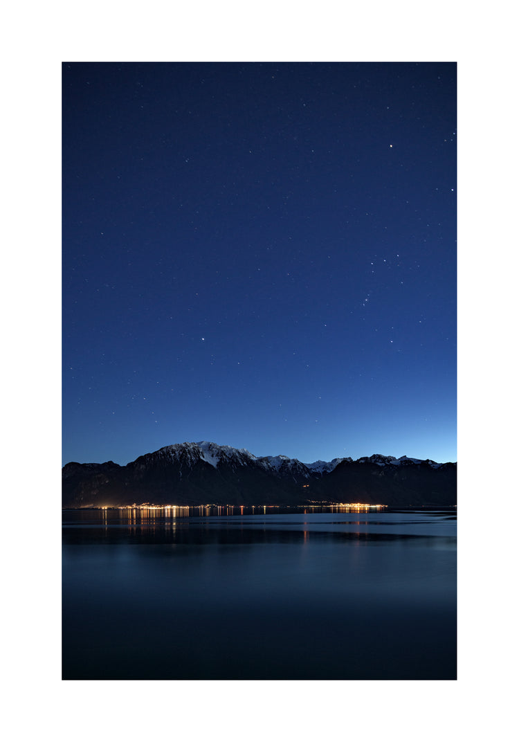 Mountain scenery, lights from a city and a lake under a star strewn sky.