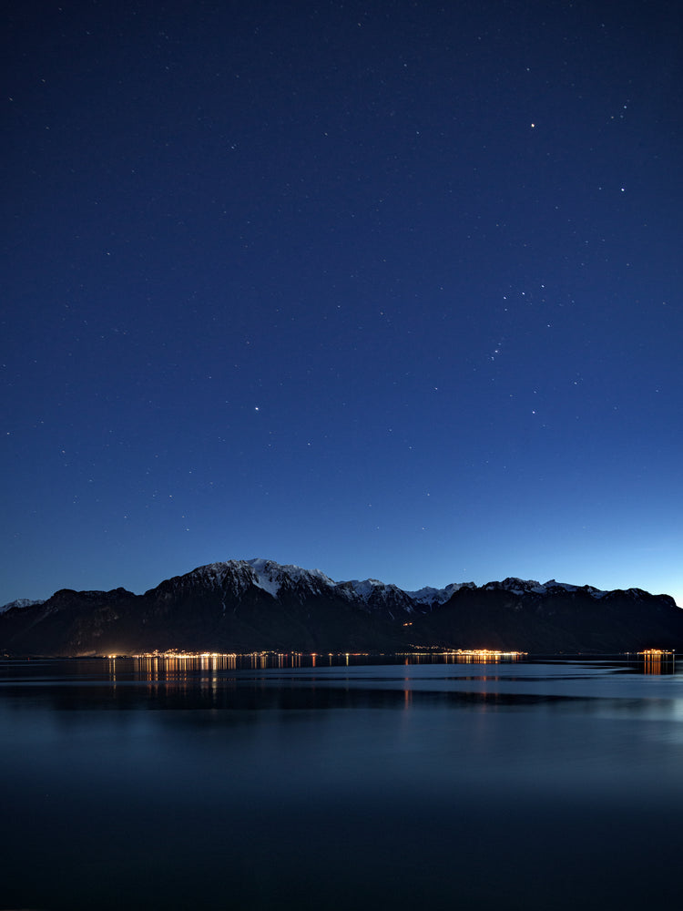 Mountain scenery, lights from a city and a lake under a star strewn sky.