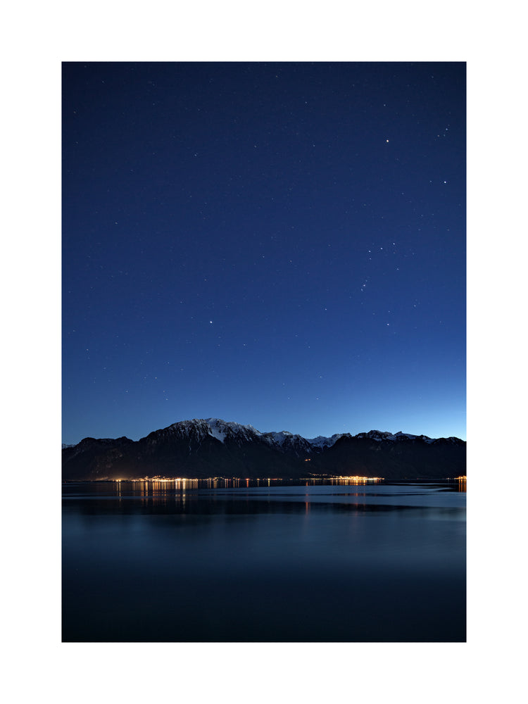 Mountain scenery, lights from a city and a lake under a star strewn sky.