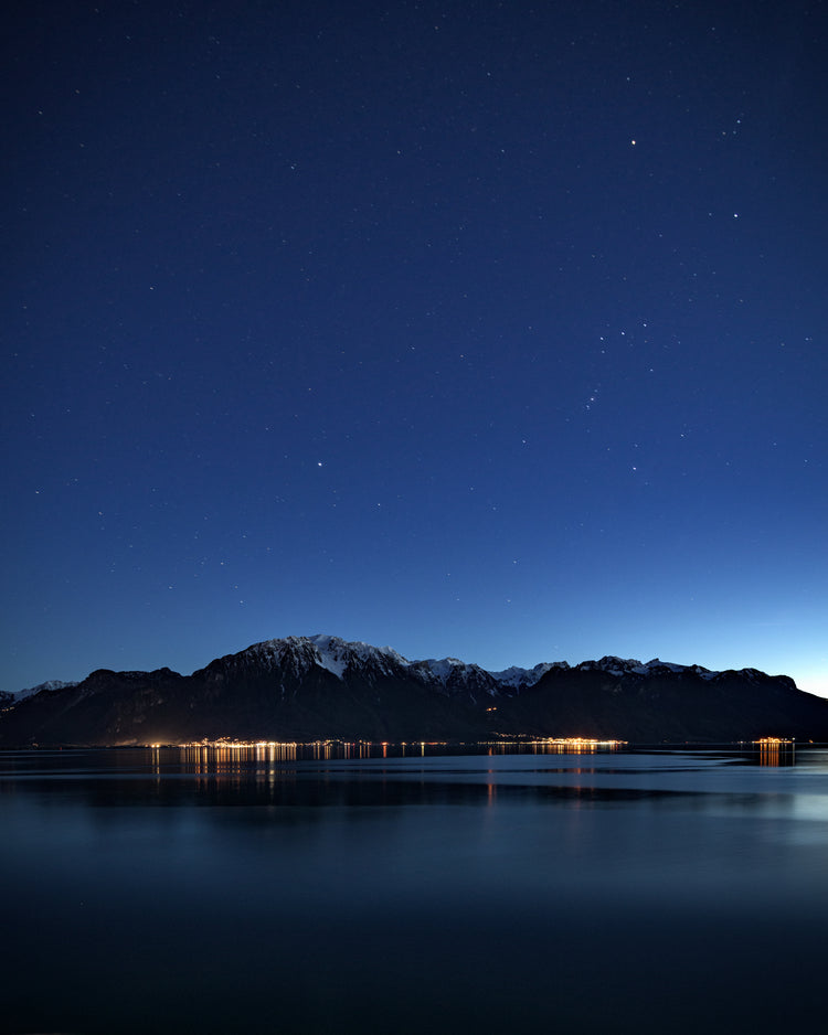 Mountain scenery, lights from a city and a lake under a star strewn sky.