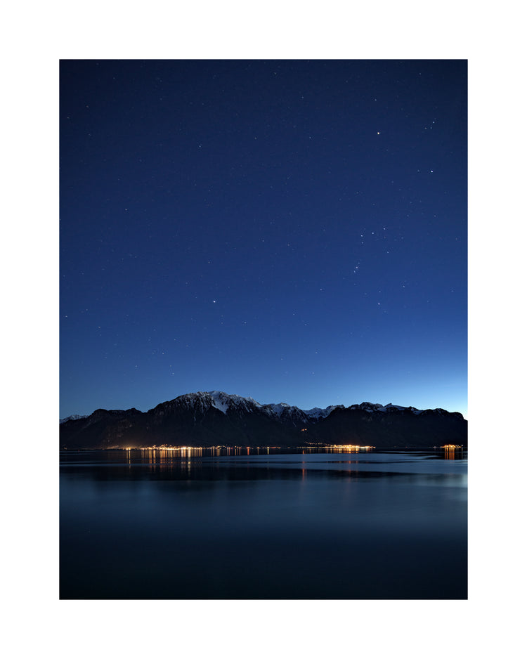Mountain scenery, lights from a city and a lake under a star strewn sky.