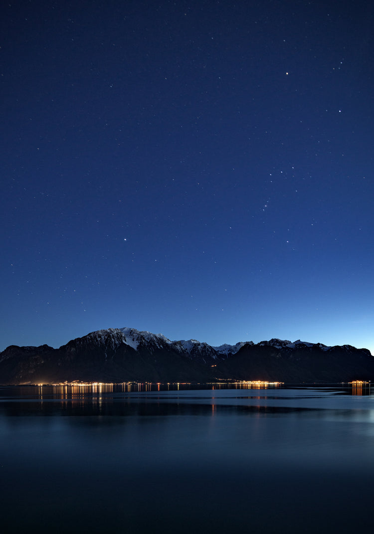 Mountain scenery, lights from a city and a lake under a star strewn sky.