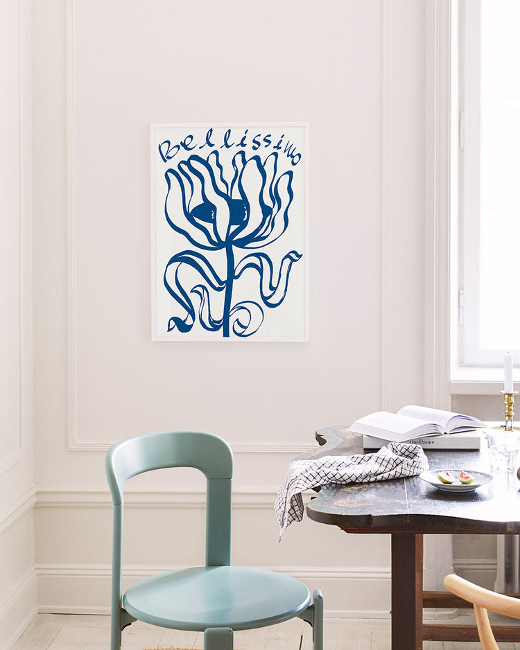A minimalist room features a light green chair and a wooden table with an open book and checkered cloth. The wall displays the Bellissimo Poster by Manne Jalilian, showcasing blue abstract flowers. The space is bright and airy with white walls and trim.