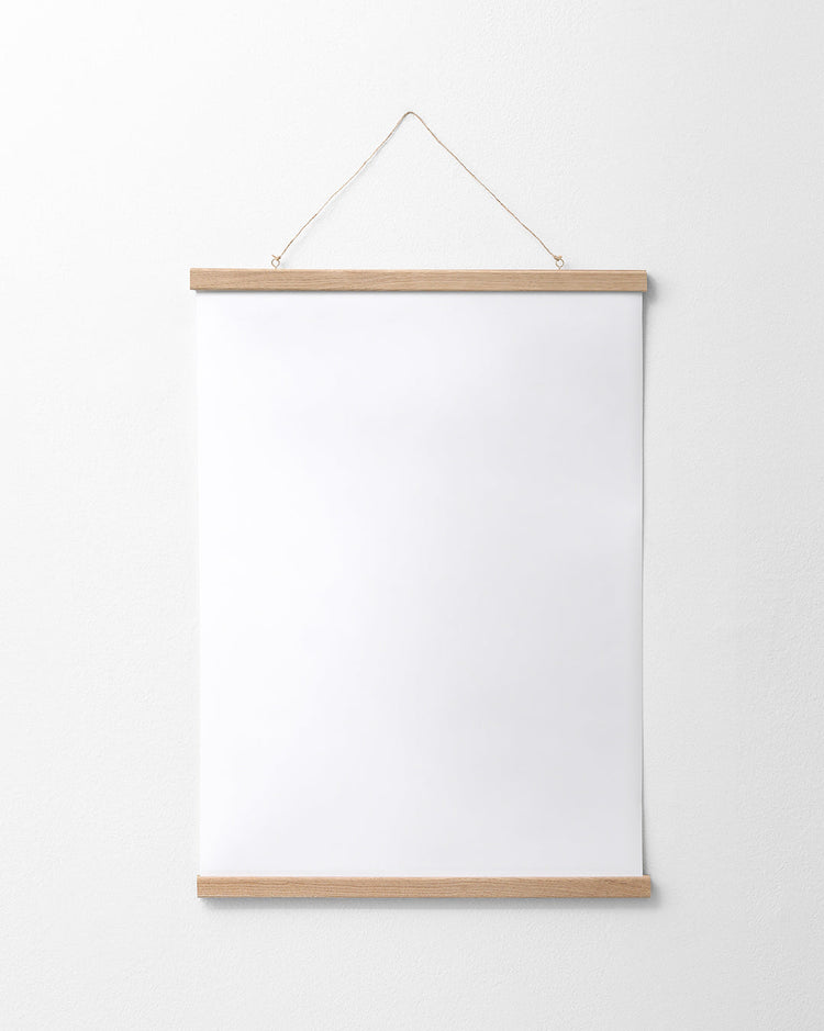A blank white poster is elegantly displayed on a white wall, framed with the Poster hanger Oak at the top and bottom. It is suspended by a thin attached string, creating a seamless look that highlights its simple design.