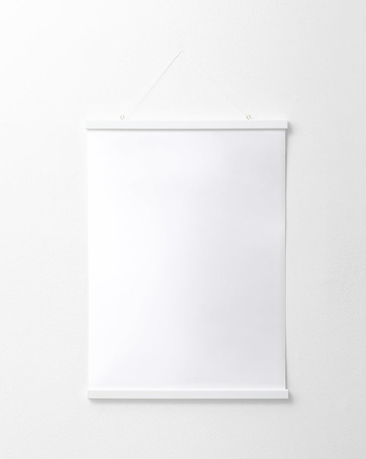 The Poster hanger White elegantly displays a blank white poster on a white wall. It features thin solid pine strips at the top and bottom, suspended by a string from two small nails, enhancing its minimalist charm.