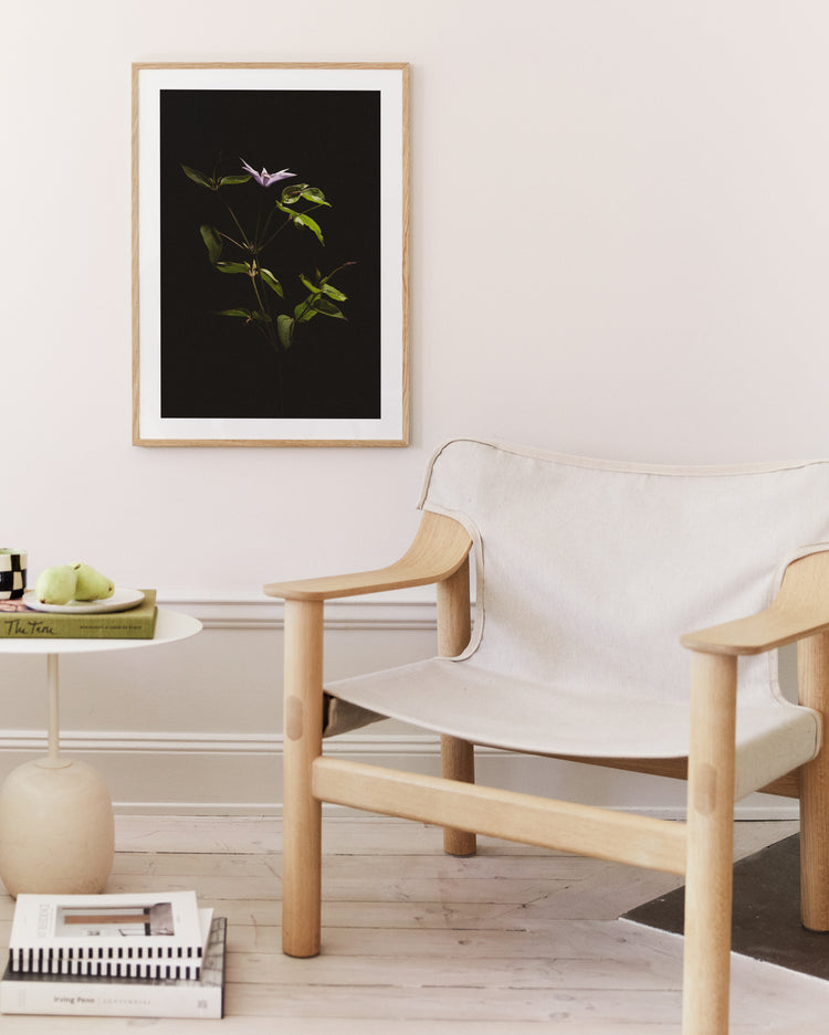 A minimalist living room features a light wood chair and a chic round side table with a book and green apples. A framed If Monet Had a Camera II poster adorns the wall, creating an atmosphere that feels both calm and elegant, much like the effortless style of French girls.