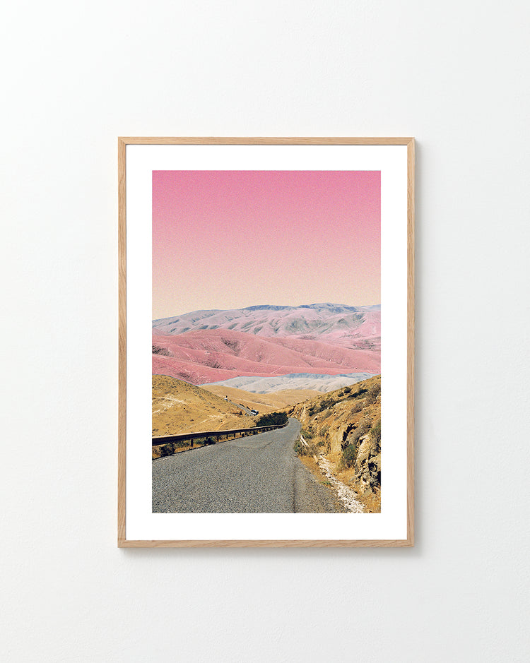 The Rosé Road Poster features a framed photograph of a winding road through hills in earthy tones under a surreal pink sky. The light wood frame stands out against a plain white wall.