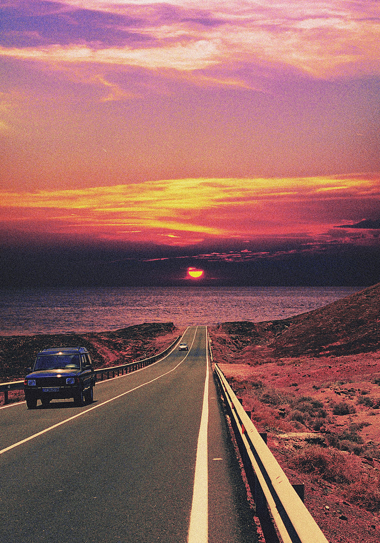 Road winding its way towards the ocean where a sun is setting on the horizon.