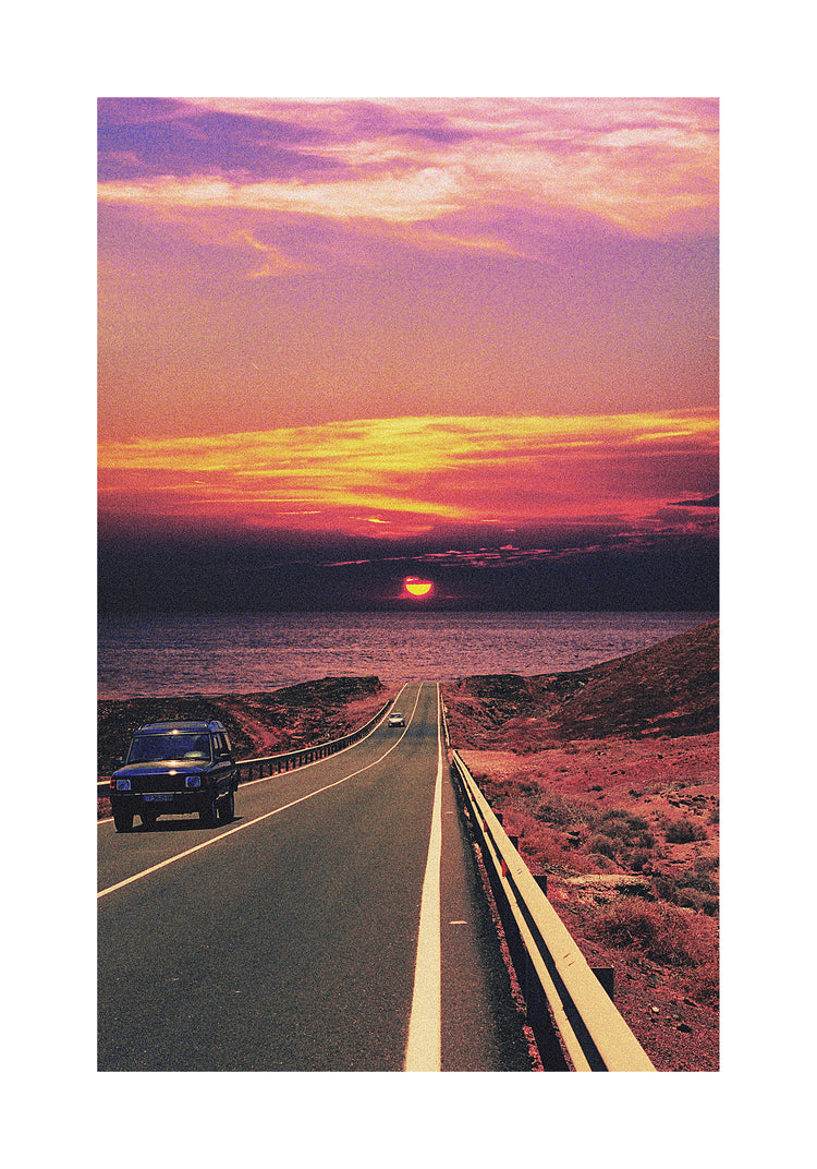 Road winding its way towards the ocean where a sun is setting on the horizon.
