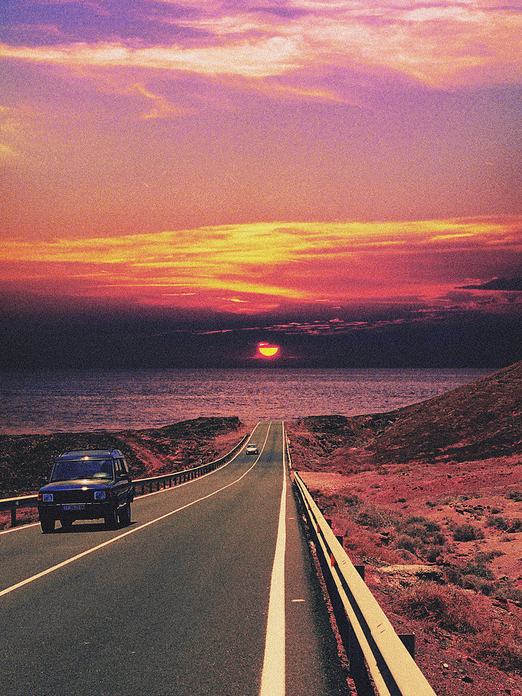 Road winding its way towards the ocean where a sun is setting on the horizon.