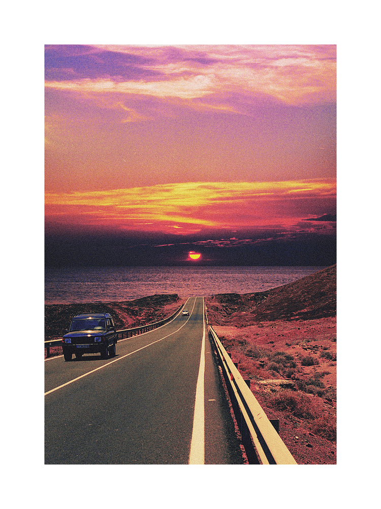 Road winding its way towards the ocean where a sun is setting on the horizon.