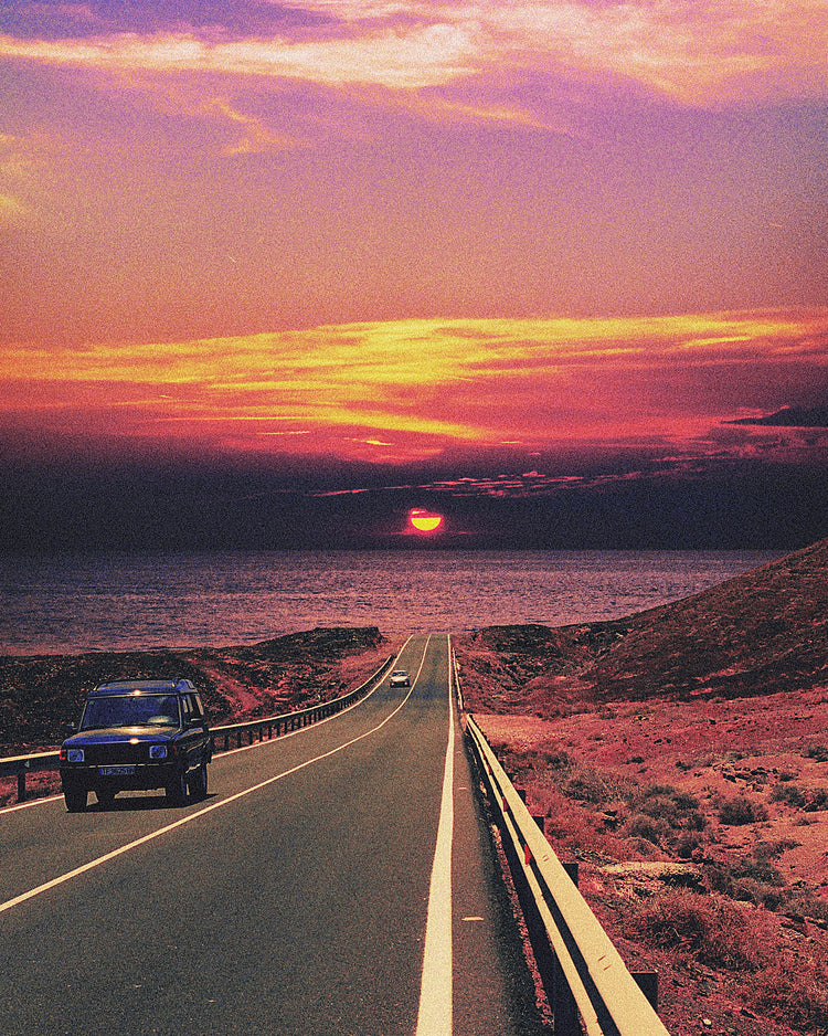 Road winding its way towards the ocean where a sun is setting on the horizon.
