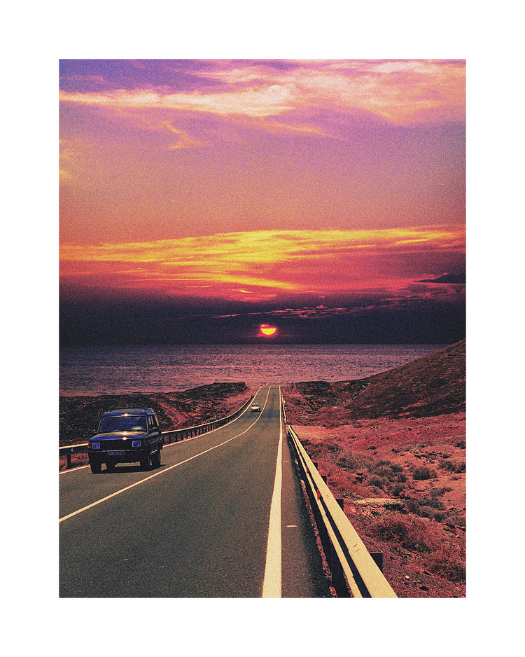 Road winding its way towards the ocean where a sun is setting on the horizon.