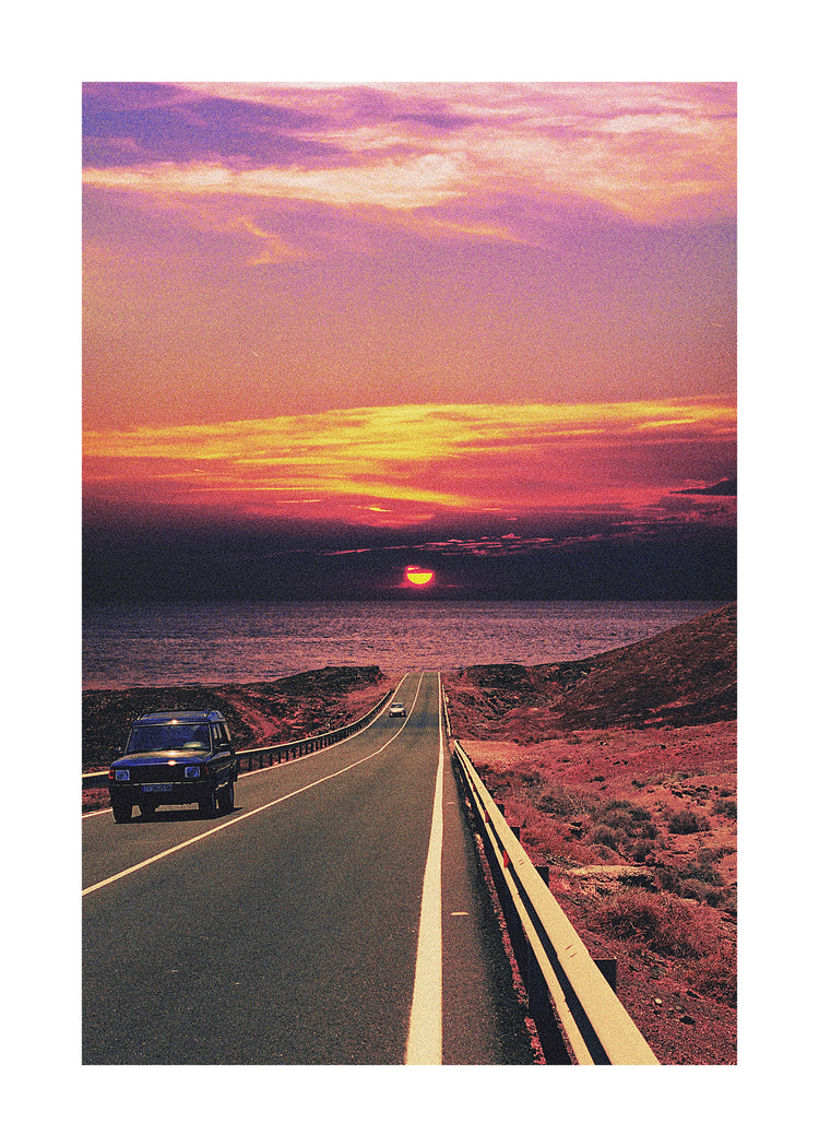 Road winding its way towards the ocean where a sun is setting on the horizon.