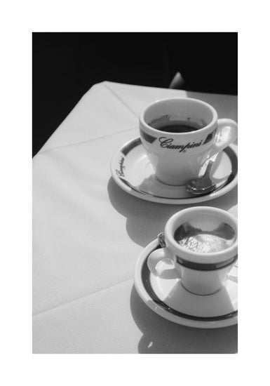 The Ciampini Poster features black and white photography capturing two espresso cups on a white tablecloth, each with a saucer and spoon. The cursive writing evokes vintage Romes charm, creating a classic ambiance with sunlight casting soft shadows.