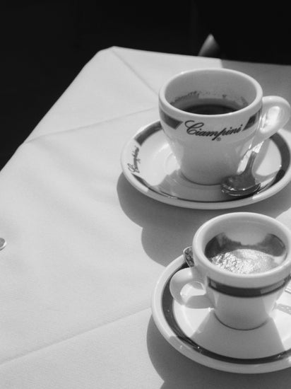 Two espresso cups on saucers with spoons sit on a white tablecloth. Emblazoned with Ciampini, they evoke vintage Romes charm, while the black and white photography enhances shadows and contrasts, reminiscent of an elegant Ciampini Poster.