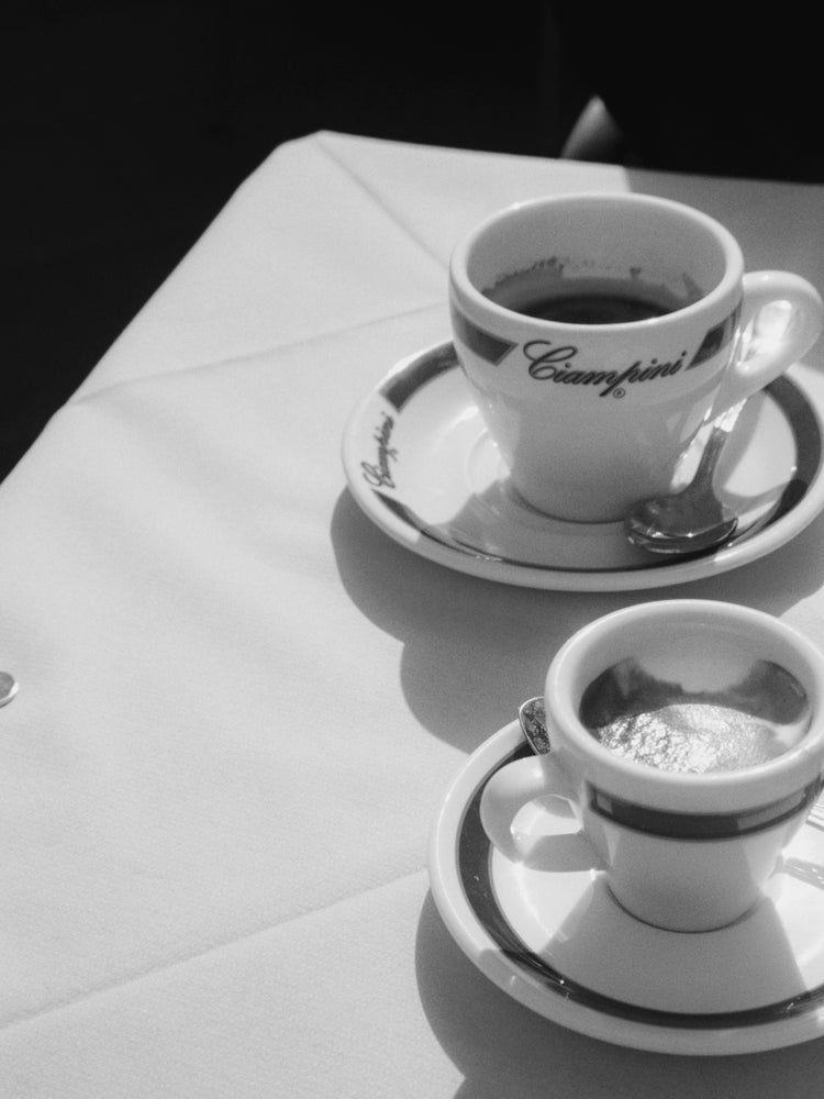Two espresso cups on saucers with spoons sit on a white tablecloth. Emblazoned with Ciampini, they evoke vintage Romes charm, while the black and white photography enhances shadows and contrasts, reminiscent of an elegant Ciampini Poster.