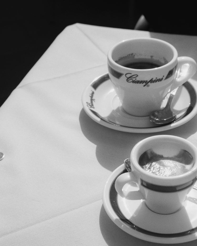 Two cups of coffee rest elegantly on a white tablecloth in the Ciampini Poster. The larger labeled cup sits on a saucer with spoon, evoking vintage Roman charm, while the smaller espresso cup accompanies it. This black-and-white photography casts soft shadows across the scene.