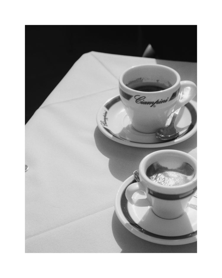 A black and white photo of two coffee cups on a sunlit table captures the charm of vintage Rome. The larger cup and an espresso, both on saucers with spoons, rest on a cloth that highlights shadows, reminiscent of a classic Ciampini Poster.
